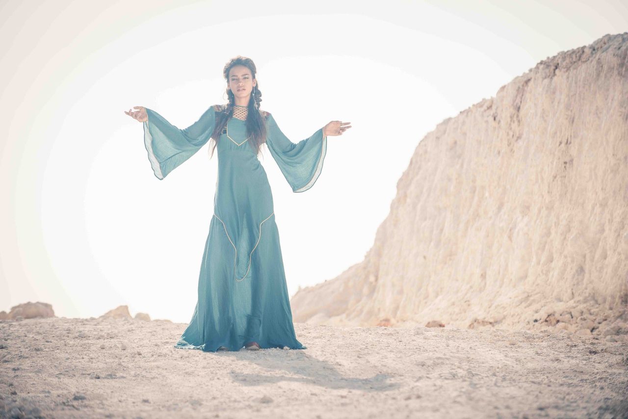 Woman in a long teal dress with flared sleeves standing on a rocky hillside
