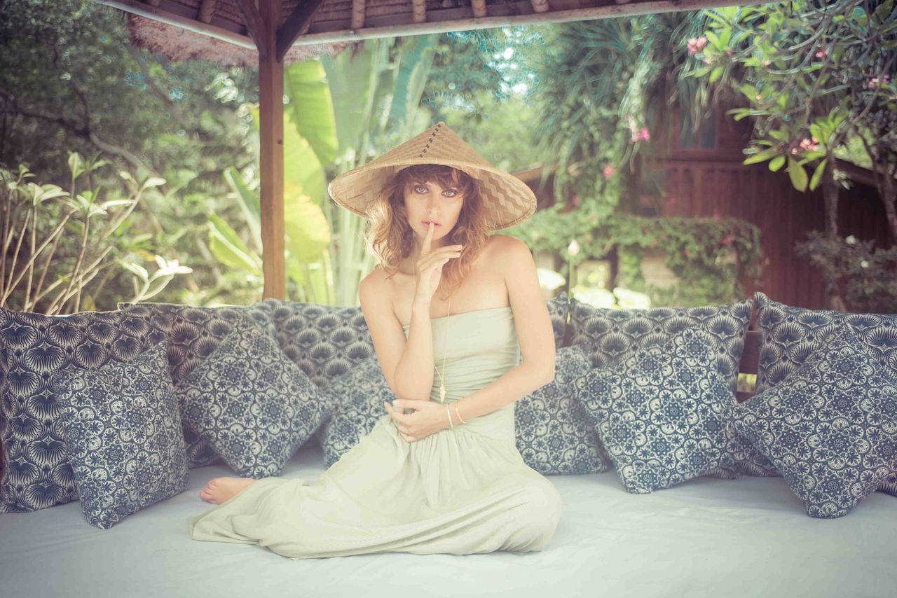 Woman in a straw hat and pale dress seated on patterned cushions in a garden