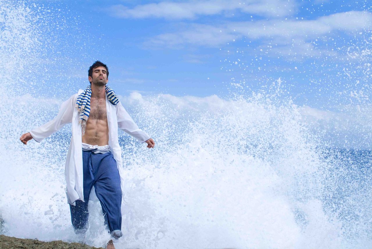 Man in an open white shirt as a wave crashes behind him