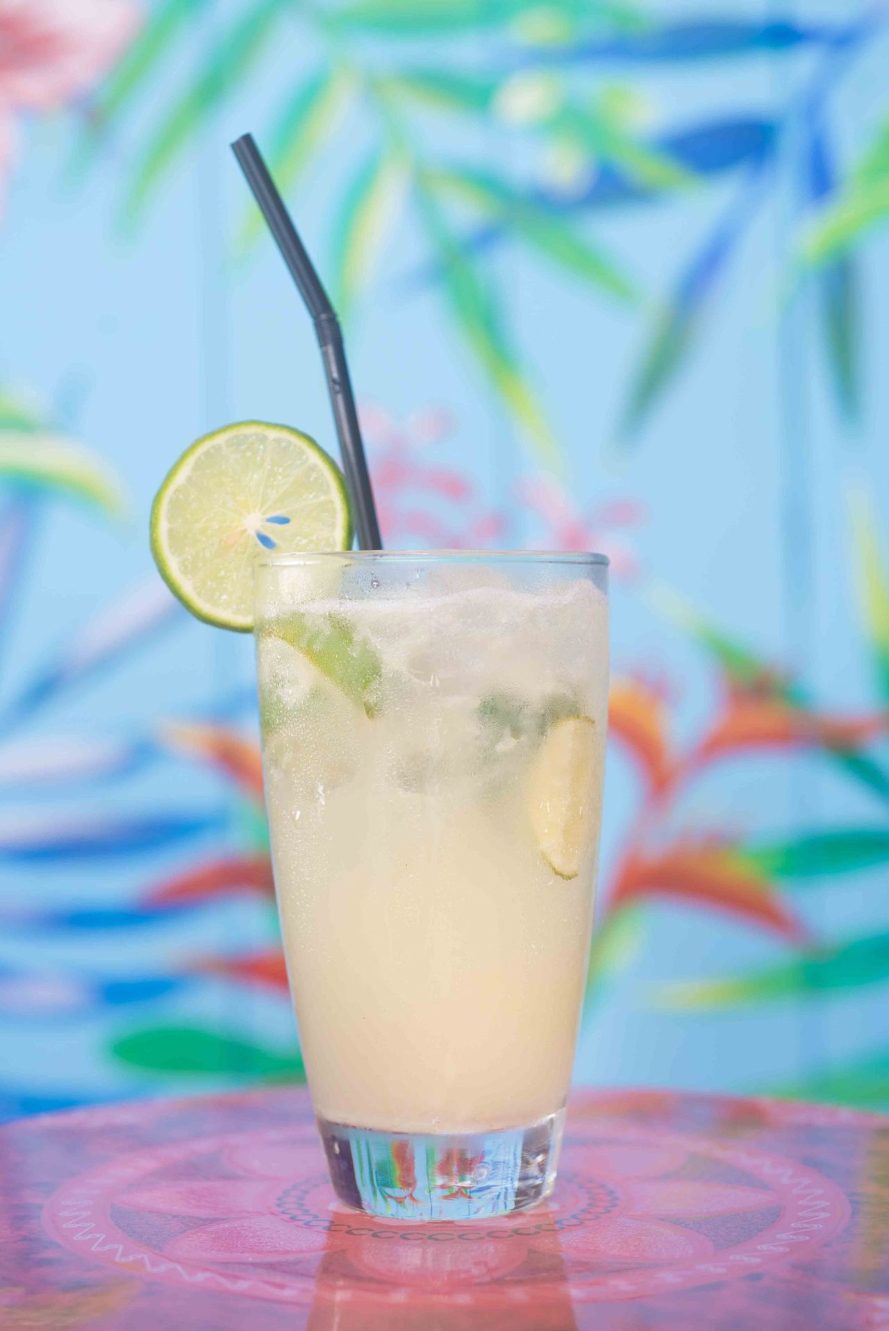 Pale drink with a lime slice and black straw in front of colourful artwork