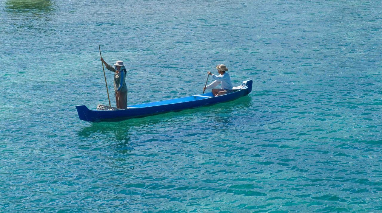 Fisherman standing in a blue outrigger canoe on clear turquoise water
